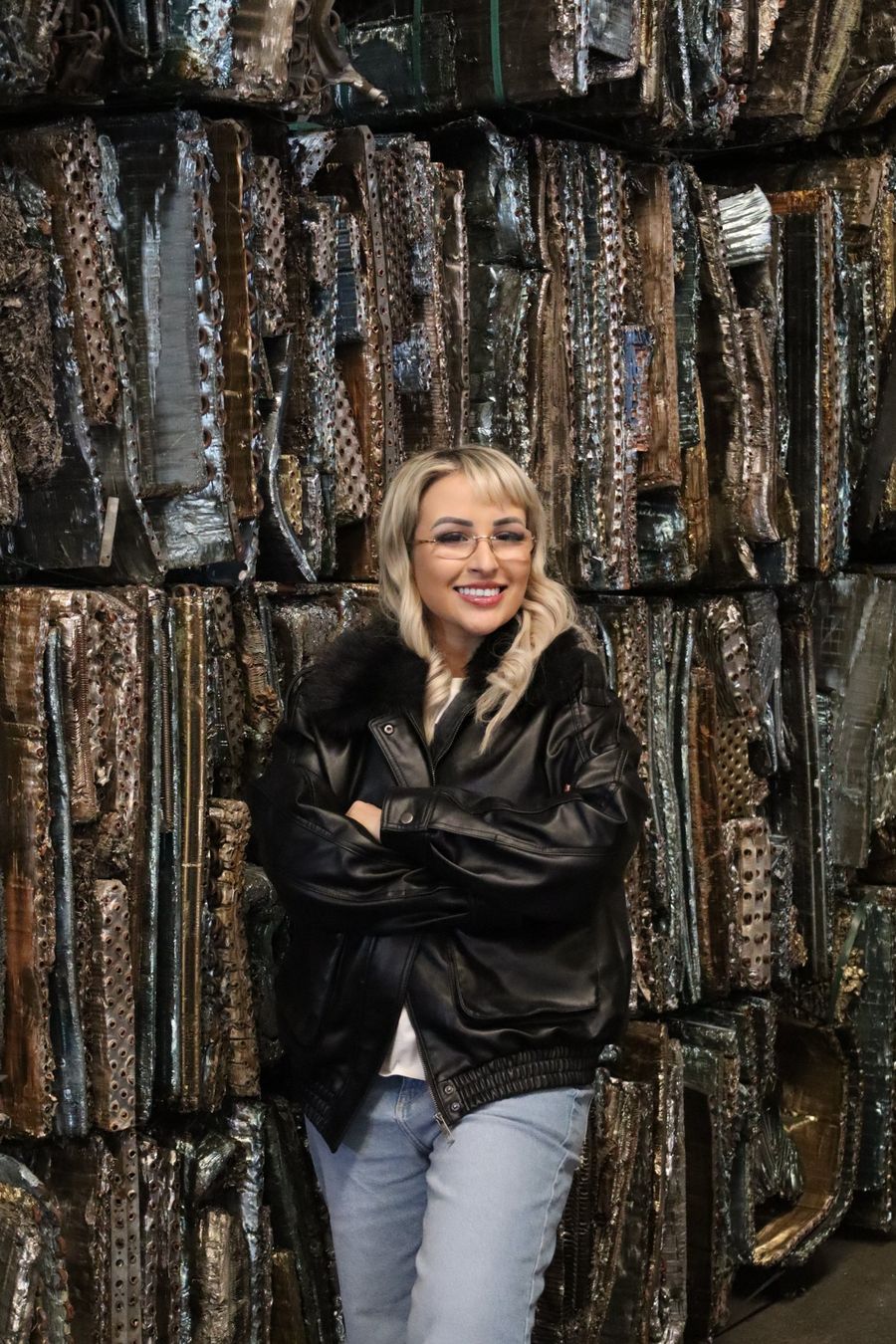 Rita Sucatas, de jaqueta preta e braços cruzados, em frente a fardos de sucata metálica